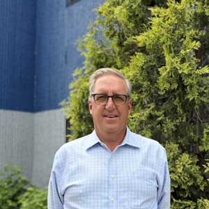 Man in glasses standing by green foliage and blue wall